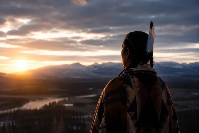 An Indigenous person wrapped in a patterned blanket, a feather in their braided hair, looking out over a river valley at sunset